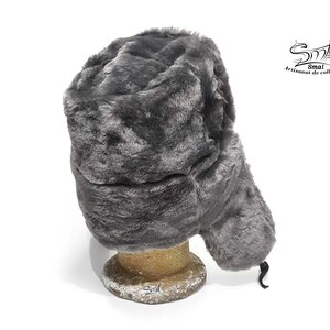 Op de afbeelding: Een grijze imitatiebont ushanka-muts met een oorflap die onder de kin kan worden vastgebonden. De muts staat op een houten standaard.