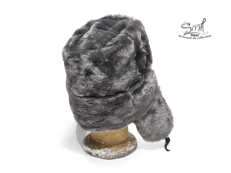 Op de afbeelding: Een grijze imitatiebont ushanka-muts met een oorflap die onder de kin kan worden vastgebonden. De muts staat op een houten standaard.