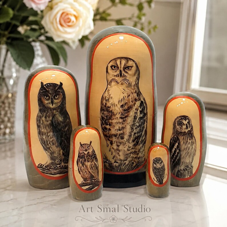 Owls hand-painted on five Russian nesting dolls, with each doll featuring a different owl in detailed natural brown, beige, and gray plumage. The set is displayed on a light marble surface in a bright elegant interior, with white roses and green foliage in a vase on the left and a sunlit window with soft curtains in the background