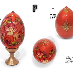 Œuf de Pâques orange intense au reflet nacré, décor floral peint à la main dans le style russe, rehaussé d’ornements dorés et d’un cabochon central, présenté avec socle, hauteur totale 12,5 cm, hauteur de l’œuf 9 cm