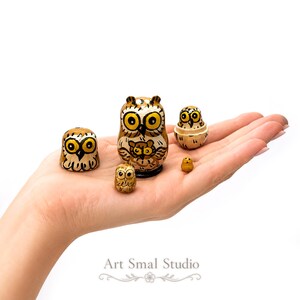 Nestled in the palm of a hand, a five-piece owl nesting doll set reveals its tiny graduated figures, including the smallest owl inside, highlighting the miniature scale of this Russian matryoshka; the set is photographed against a bright white background