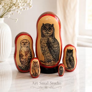 Hand-painted owls on a set of five Russian nesting dolls, featuring several different owl species and detailed plumage in natural brown and gray tones. The Matryoshkas are arranged on a light marble surface in a bright elegant interior, with a white vase of delicate flowers and soft curtains in the background