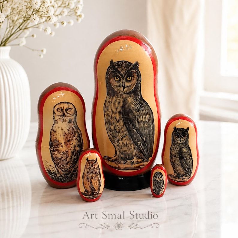 Hand-painted owls on a set of five Russian nesting dolls, featuring several different owl species and detailed plumage in natural brown and gray tones. The Matryoshkas are arranged on a light marble surface in a bright elegant interior, with a white vase of delicate flowers and soft curtains in the background