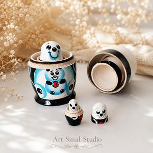 Panda matryoshka nesting dolls shown partially disassembled on a light tabletop, with the largest doll open and its inner wooden layers visible. Smaller nesting doll pieces are arranged in front, while softly blurred dried flowers and natural fabric create a warm, elegant background