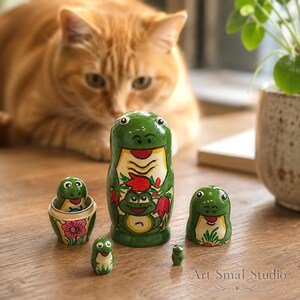 A playful frog nesting doll collection sits on a wooden tabletop while a softly blurred ginger cat curiously watches from behind, creating a charming animal-themed home décor scene