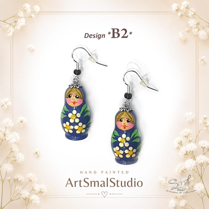 Hand-painted blue matryoshka doll earrings – Design B2. Adorable royal blue Russian nesting doll earrings decorated with white and yellow floral patterns, green leaf accents, rosy cheeks, and blonde hair details. Lightweight folk-art inspired dangle earrings with silver-tone hooks and elegant handcrafted charm by ArtSmalStudio
