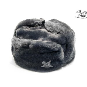 Op de afbeelding: Een grijze Ushanka-muts van imitatiebont met een omklapbare oorflap. De muts heeft een klein geborduurd logo met de tekst "Smal Artisanat de collection".