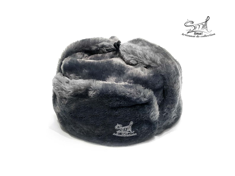 Op de afbeelding: Een grijze Ushanka-muts van imitatiebont met een omklapbare oorflap. De muts heeft een klein geborduurd logo met de tekst "Smal Artisanat de collection".