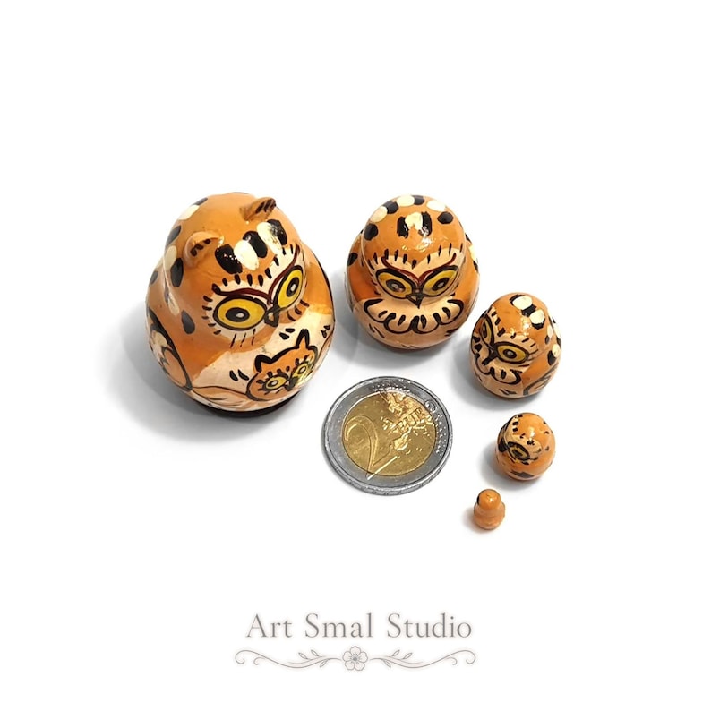 A charming owl matryoshka nesting doll set with five hand-painted wooden owls in warm orange and cream tones, decorated with expressive golden eyes, folk-art details, and delicate owl motifs. The smallest pieces are displayed beside a 2 euro coin to show their remarkably tiny scale, highlighting this miniature Russian nesting doll design and its intricate craftsmanship. Presented on a clean white background with soft shadows
