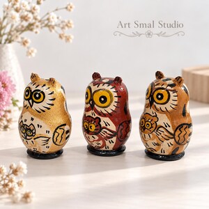 Three hand-painted owl matryoshka nesting dolls in gold, burgundy and warm brown tones, featuring expressive golden eyes and detailed folk-art designs. Displayed on a light wooden surface with delicate white flowers and a soft neutral background