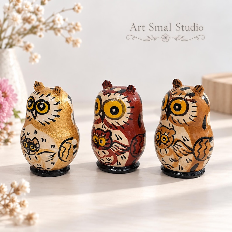Three hand-painted owl matryoshka nesting dolls in gold, burgundy and warm brown tones, featuring expressive golden eyes and detailed folk-art designs. Displayed on a light wooden surface with delicate white flowers and a soft neutral background