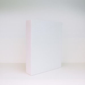 May include: A white rectangular block with a textured surface and a pink edge, positioned on a white shelf against a white background. The object is a simple, geometric shape, likely used for display or product photography.