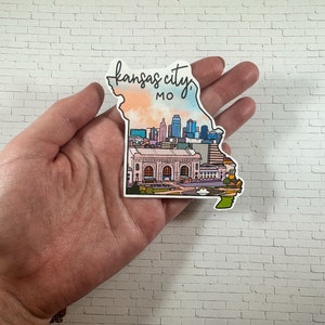 Puede incluir: Una pegatina que representa el horizonte de Kansas City, Misuri. La pegatina tiene la forma del estado de Misuri y presenta una ilustraci&oacute;n colorida del horizonte de la ciudad, incluida la estaci&oacute;n Union. El texto "Kansas City, MO" est&aacute; escrito en cursiva en la parte superior de la pegatina.