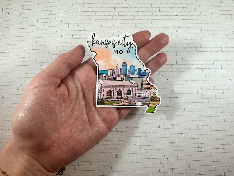 Puede incluir: Una pegatina que representa el horizonte de Kansas City, Misuri. La pegatina tiene la forma del estado de Misuri y presenta una ilustraci&oacute;n colorida del horizonte de la ciudad, incluida la estaci&oacute;n Union. El texto "Kansas City, MO" est&aacute; escrito en cursiva en la parte superior de la pegatina.