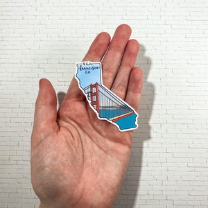 Op de afbeelding: Een sticker van de staat Californi&euml; met de Golden Gate Bridge in de vorm van de staat. De sticker is blauw, rood en zwart met de tekst "San Francisco, CA" bovenaan.