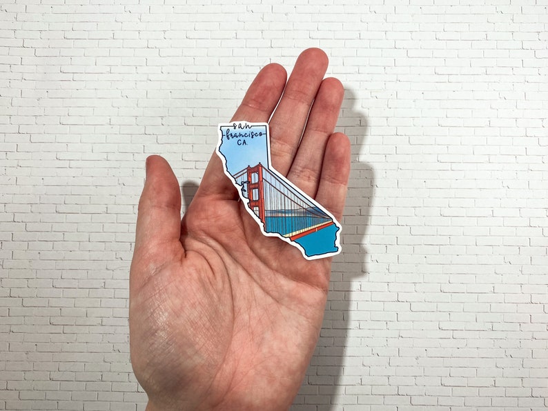 Op de afbeelding: Een sticker van de staat Californi&euml; met de Golden Gate Bridge in de vorm van de staat. De sticker is blauw, rood en zwart met de tekst "San Francisco, CA" bovenaan.