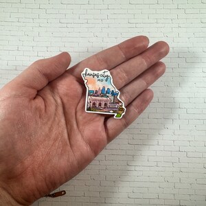 Puede incluir: Una pegatina con el horizonte de Kansas City en forma del estado de Misuri. La pegatina es una ilustraci&oacute;n de acuarela con un esquema de color rosa, naranja y azul. El texto "Kansas City, MO" est&aacute; escrito en cursiva negra en la parte superior de la pegatina.
