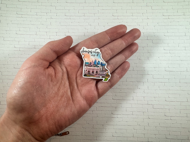 Puede incluir: Una pegatina con el horizonte de Kansas City en forma del estado de Misuri. La pegatina es una ilustraci&oacute;n de acuarela con un esquema de color rosa, naranja y azul. El texto "Kansas City, MO" est&aacute; escrito en cursiva negra en la parte superior de la pegatina.