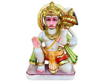 May include: A white marble statue of the Hindu deity Hanuman, sitting with his right hand raised in a gesture of blessing. He is holding a golden mace in his left hand. The statue is painted with gold, red, and green accents.