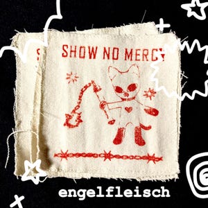 May include: Off-white fabric patch with red print. The text "SHOW NO MERCY" is above a cartoon cat-like figure holding a flail. Below the figure is the word "engelfleisch". The patch has a raw, unfinished edge.