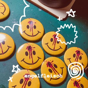 May include: Yellow smiley face buttons with a black smile and a red blood splatter design. The buttons have the word "engelfleisch" printed on them. The buttons are arranged on a green surface.