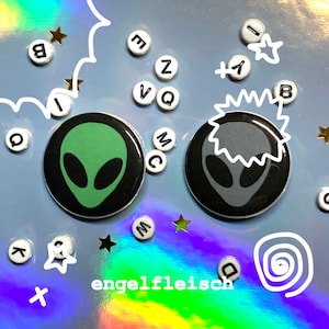 May include: Two circular black badges featuring alien faces, one green and one grey, are shown on a holographic surface. White letter beads and star-shaped confetti surround the badges. The word "engelfleisch" is at the bottom.