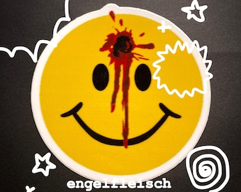 May include: A yellow smiley face sticker with a bullet hole and blood splatter graphic. The sticker has a white border and the word "engelfleisch" is printed at the bottom. Decorative white doodles surround the smiley face.