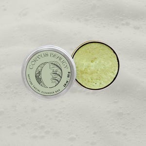 May include: A silver tin of Corpus Beauty Neptune Facial Cleanser Bar, with a light green label featuring a crescent moon and leaf design. Beside it, the open tin reveals a light green, textured cleanser bar, weighing 28 g / 1 oz.