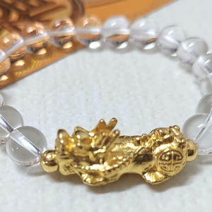 May include: A clear crystal beaded bracelet with a gold-colored dragon charm. The charm has intricate details and a Chinese character on its side.