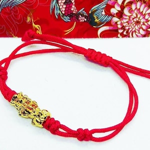 May include: A red string bracelet with a gold Chinese lucky charm. The charm is a detailed depiction of a mythical creature.