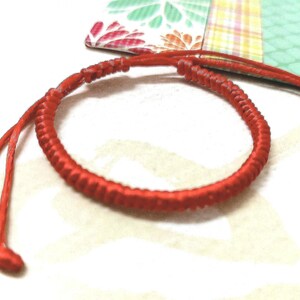 Puede incluir: Un brazalete de cuerda roja con un diseño anudado. El brazalete está atado con un nudo simple en el extremo.