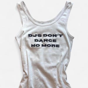 Può includere: Canotta bianca con la scritta "DJs Don't Dance No More" in grassetto nero. La canotta ha uno scollo tondo e un design senza maniche. Il tessuto sembra essere un materiale morbido e leggero.