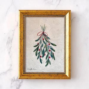 Op de afbeelding: Een ingelijst schilderij met een aquarelillustratie van een maretak met groene bladeren en rode bessen, samengebonden met een dun rood lint. Het kunstwerk zit in een rechthoekige gouden lijst met een gedetailleerde, kralen binnenrand.