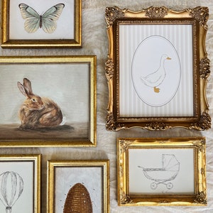 May include: Five gold picture frames with different images inside. The top frame has a white butterfly on a light brown background. The frame to the right has a white duck on a light gray striped background. The frame below the butterfly has a brown rabbit on a light brown background. The frame below the duck has a black and white illustration of a baby carriage on a white background. The frame to the right of the rabbit has a brown beehive on a light brown background.