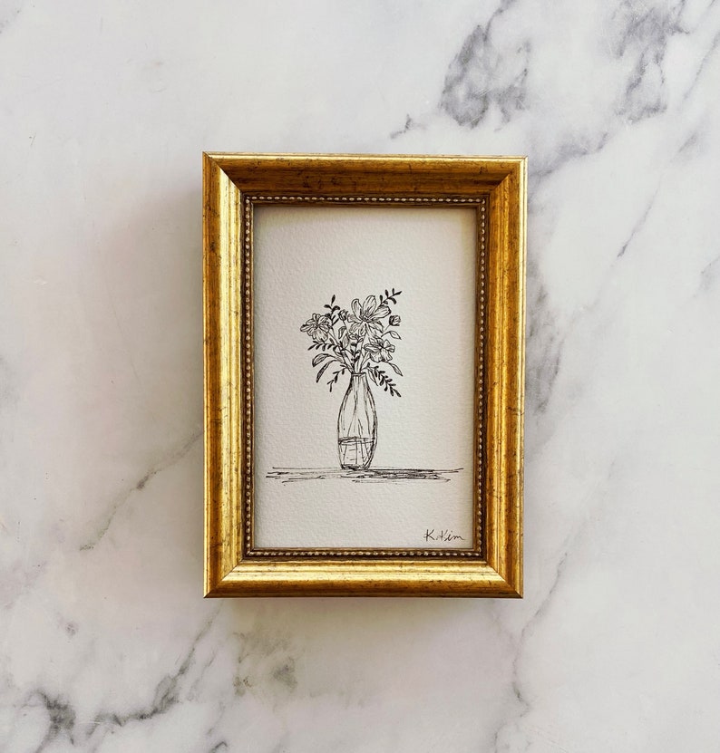 May include: A black and white line drawing of a bouquet of flowers in a vase, framed in a gold frame. The drawing is signed "Kaitlin".
