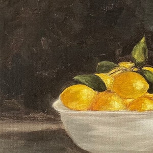 May include: A white ceramic bowl filled with yellow lemons and green leaves. The bowl is sitting on a dark brown wooden surface.