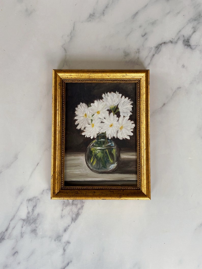 May include: A framed painting of a bouquet of white daisies in a clear glass vase. The daisies are arranged in a cluster and the vase is sitting on a table. The background is a dark brown color.