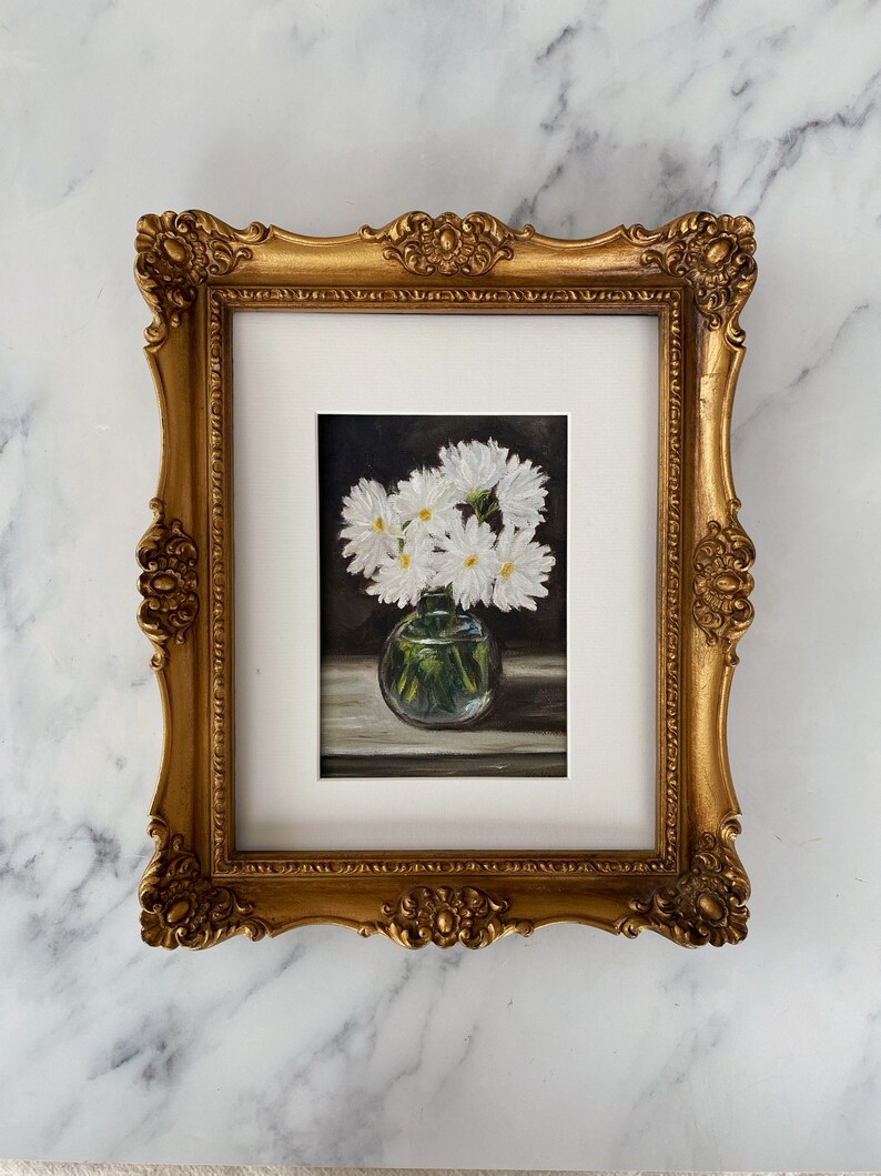 May include: A framed painting of a bouquet of white daisies in a clear glass vase. The daisies are arranged in a simple, elegant way, and the painting is done in a realistic style.