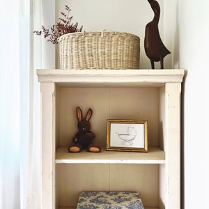 May include: A white wooden shelf with three shelves. The top shelf has a wicker basket with dried branches, a wooden duck figurine, and a white box. The middle shelf has a brown stuffed rabbit and a framed print of a baby carriage. The bottom shelf has a blue and white patterned box.