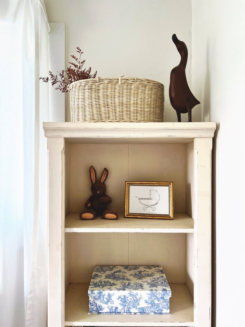 May include: A white wooden shelf with three shelves. The top shelf has a wicker basket with dried branches, a wooden duck figurine, and a white box. The middle shelf has a brown stuffed rabbit and a framed print of a baby carriage. The bottom shelf has a blue and white patterned box.