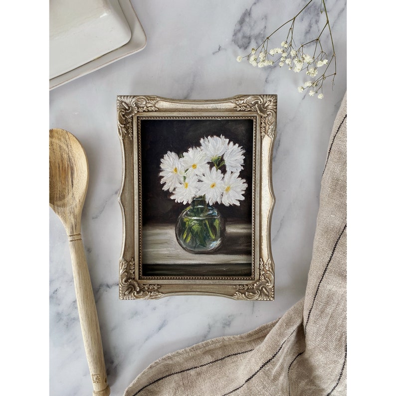 May include: A framed painting of a bouquet of white daisies in a clear glass vase. The daisies are arranged in a cluster and have a dark background.