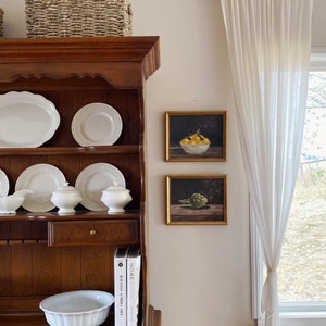 May include: A wooden hutch with white dishes, two framed still life prints, and two cookbooks titled "The Silver Spoon" and "Home Cook." The hutch is topped with a woven basket.