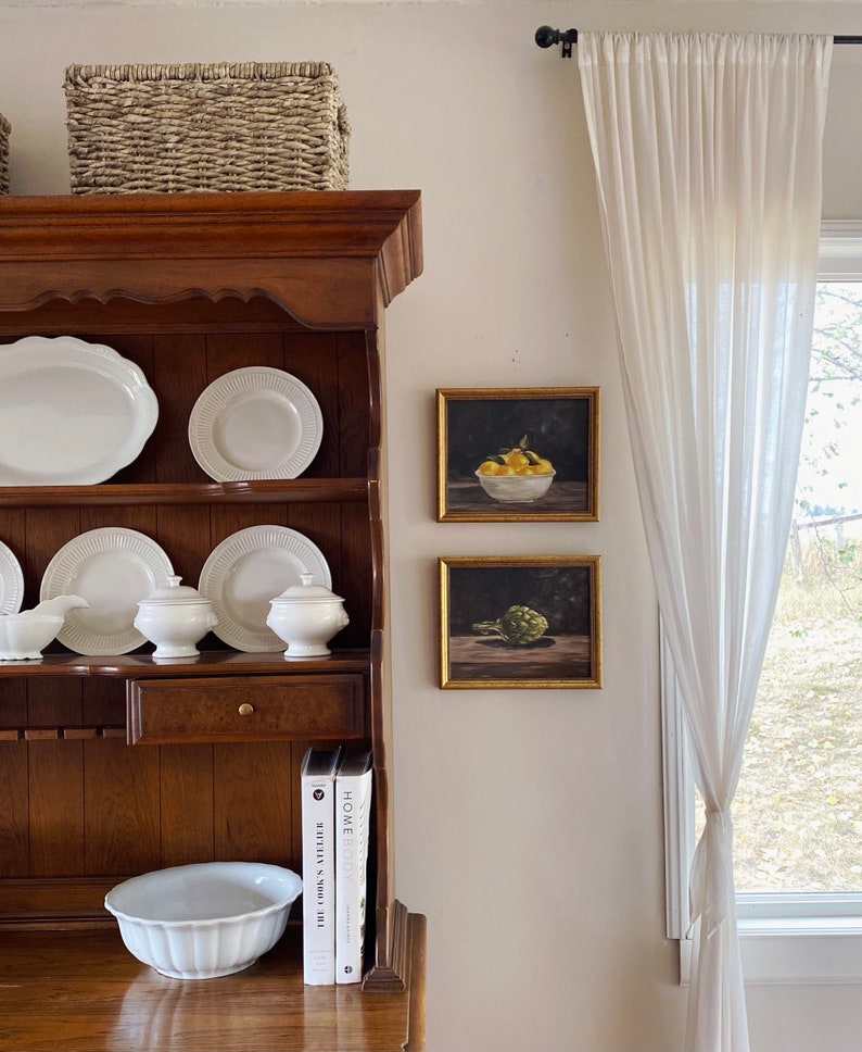 May include: A wooden hutch with white dishes, two framed still life prints, and two cookbooks titled "The Silver Spoon" and "Home Cook." The hutch is topped with a woven basket.