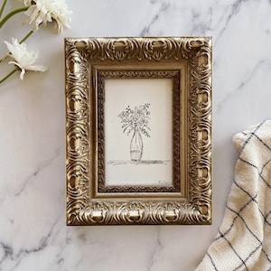 May include: A black and white line drawing of a bouquet of flowers in a vase, framed in a gold ornate frame. The drawing is signed "Kilen".