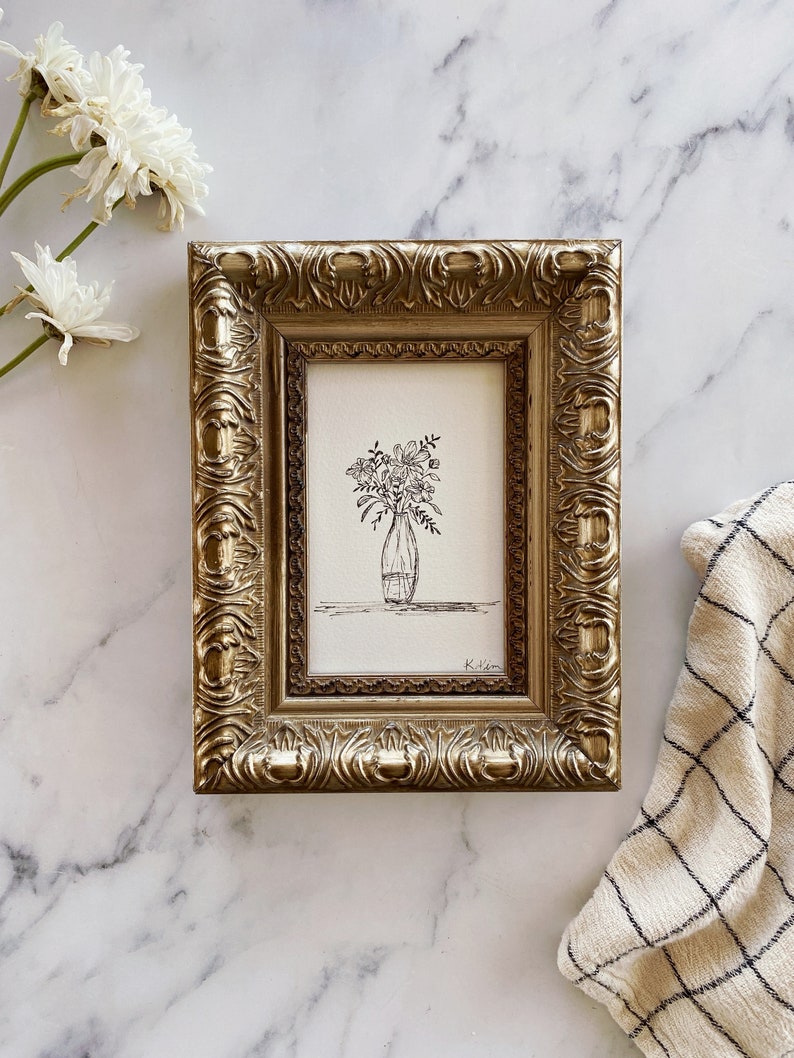 May include: A black and white line drawing of a bouquet of flowers in a vase, framed in a gold ornate frame. The drawing is signed "Kilen".