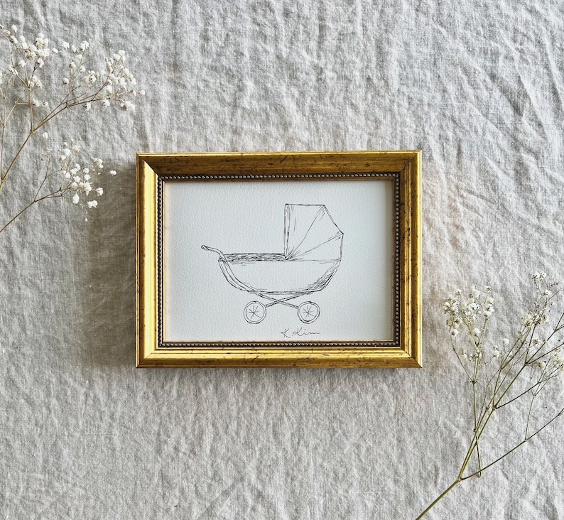 May include: A black and white line drawing of a baby carriage in a gold frame. The drawing is on white paper with a black signature in the bottom center.
