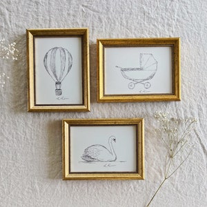 May include: Three gold picture frames with black and white line drawings inside. The frames are hanging on a white textured wall. The drawings are of a hot air balloon, a baby carriage, and a swan.