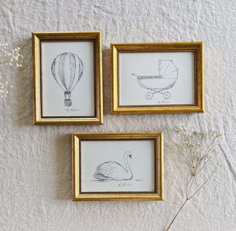 May include: Three gold picture frames with black and white line drawings inside. The frames are hanging on a white textured wall. The drawings are of a hot air balloon, a baby carriage, and a swan.