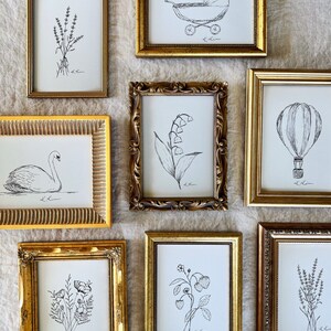 May include: A collection of eight framed black and white line drawings of various subjects, including a hot air balloon, a baby carriage, a swan, flowers, and lavender. The frames are all gold and have ornate details.