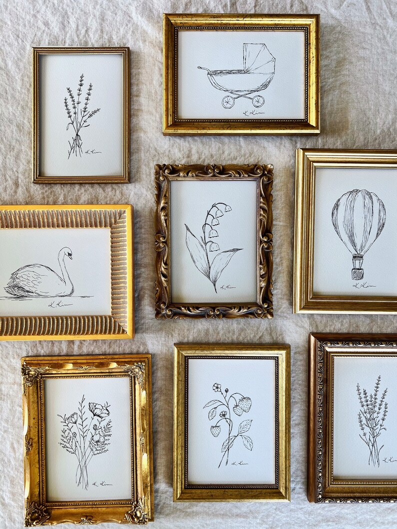 May include: A collection of eight framed black and white line drawings of various subjects, including a hot air balloon, a baby carriage, a swan, flowers, and lavender. The frames are all gold and have ornate details.